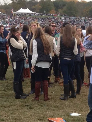 une groupes de femmes d'âge université, habillées toutes en bottes à genou, chemisier blanche ou écru, et doudounes sans manches. Elles ont toutes la même longueur de cheveux et sont dans un grand festival. On voit des milliers des personnes en fond de plan et un grand barnum. 